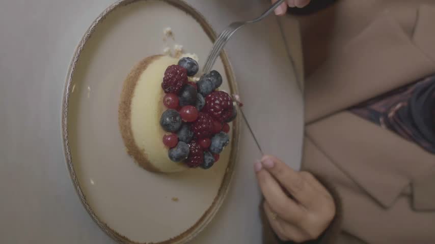 Top view of female person eating delicious cheesecake topped with fresh blueberries, raspberries, and red currants using fork and knife while sitting in restaurant.