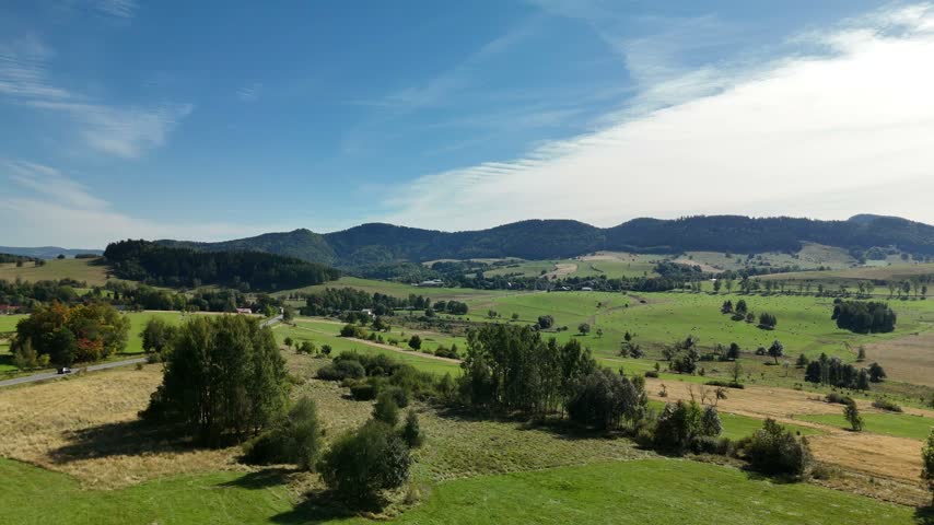Aerial view of highland meadows with scattered trees and forested mountain ridges under blue sky with wispy clouds
