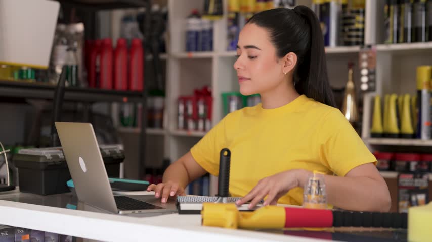 Young latina female shop assistant typing on a laptop computer behind auto parts store counter