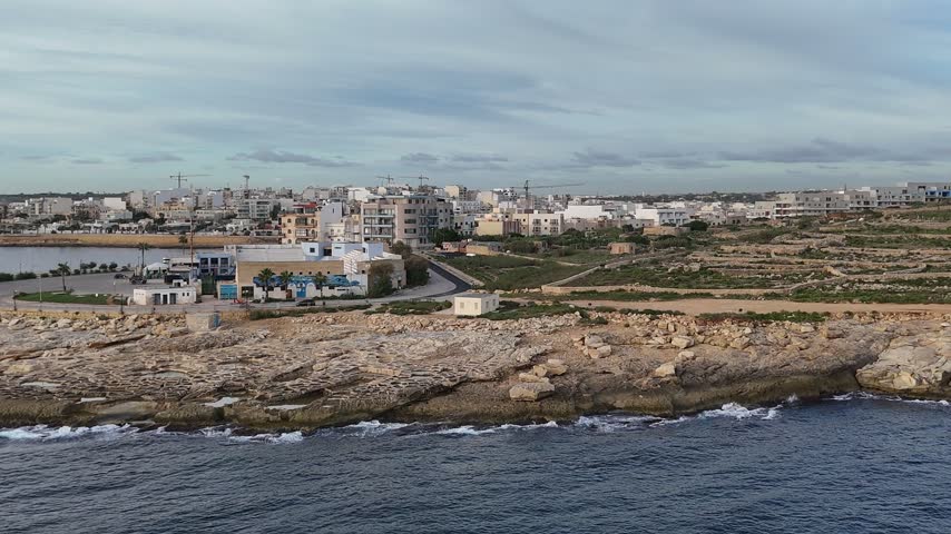 Drone rising over the shore to reveal rock-cut salt pans on a limestone point below a coastal town and bay, Malta.