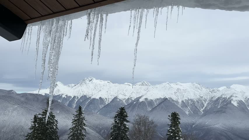 Icicles hang from a wooden roof, overlooking snow-covered mountains and evergreen trees. The scene captures a serene winter landscape.
