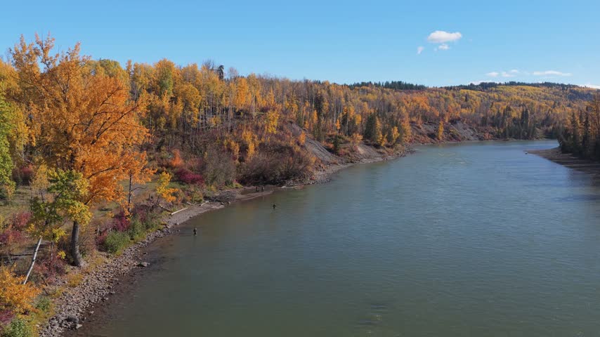 Anglers wade and fly fish near the rocky bank of a broad river bordered by golden autumn woodland and sloping hills.