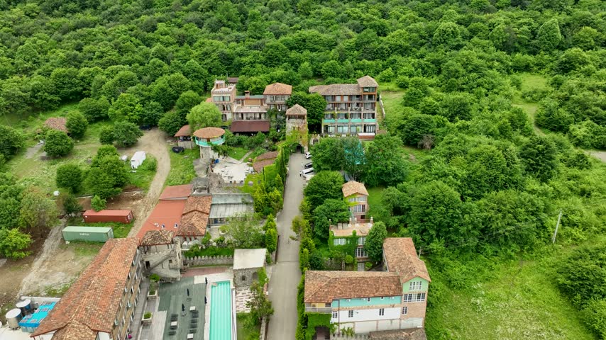 Elevated drone perspective of a small resort complex nestled within extensive green woodland in Georgia. Residential buildings, red tiled roofs, garden spaces