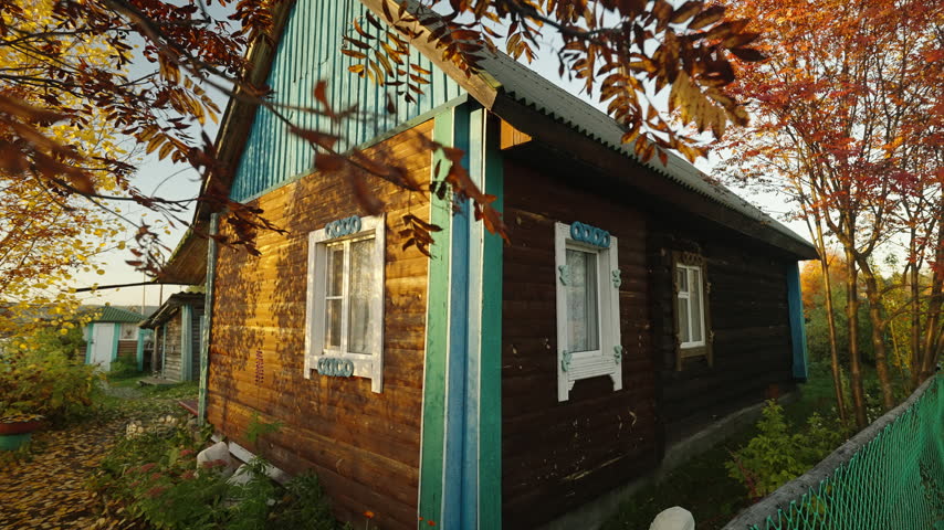 Slow pan of a traditional wooden country house with blue trim, surrounded by autumn foliage and warm sunset light in a quiet rural village. Peaceful fall countryside home exterior.