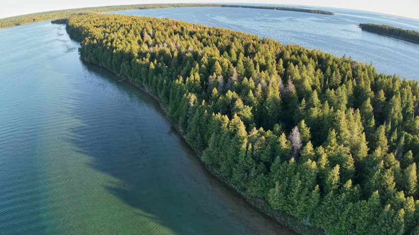 Drone view follows a long densely wooded island bordered by calm blue water in the Lake Huron region near Hessel Michigan