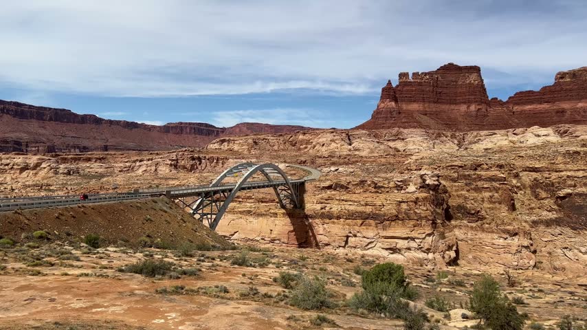 A steel arch bridge carries a winding road across a desert canyon surrounded by layered sandstone cliffs, a red butte and scattered shrubs.