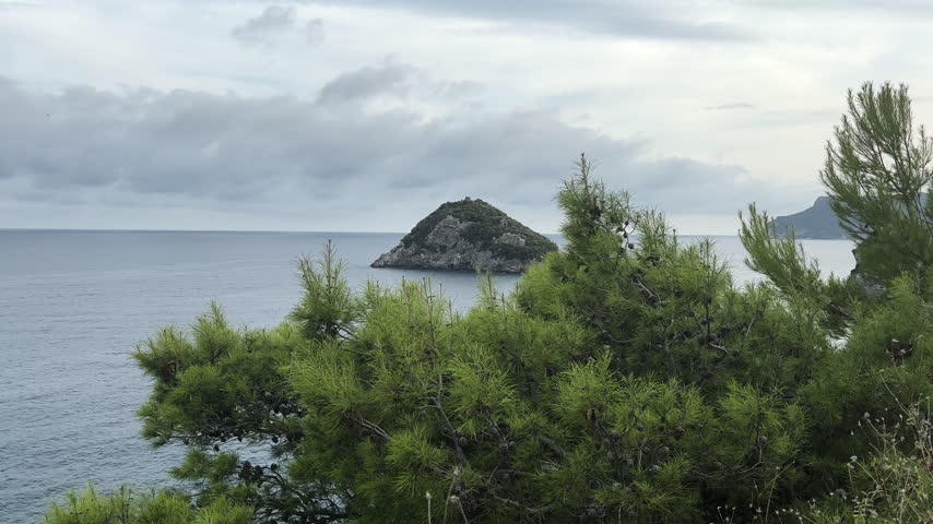 Scenic view of a small rocky island in the Ligurian Sea near Savona, Italy, framed by lush Mediterranean pine trees, calm coastal waters, distant headlands, and cloudy skies.