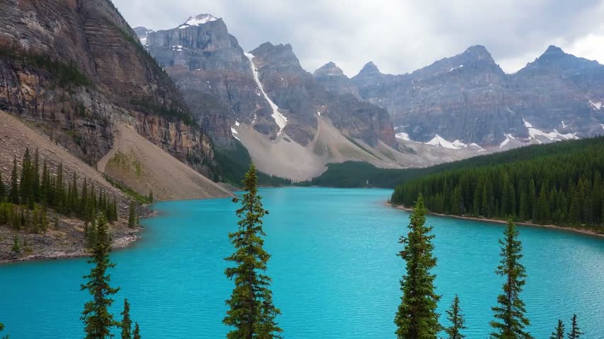 Turquoise Moraine Lake below the snow-dusted Valley of the Ten Peaks under a cloudy sky with spruce trees in the foreground, Banff National Park, Alberta, Canada
