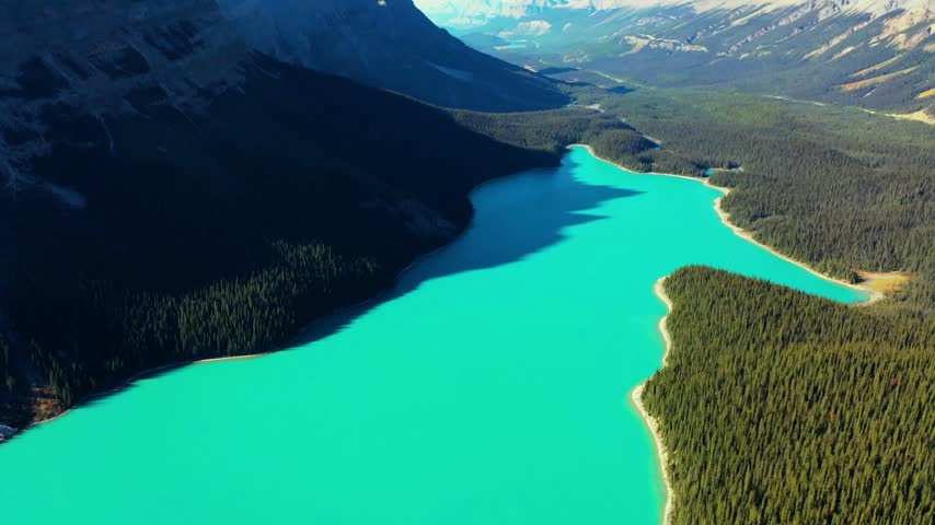 Drone view of an intensely turquoise mountain lake with a long mountain shadow, dense spruce forest and a sandy shoreline, Banff National Park, Alberta, Canada