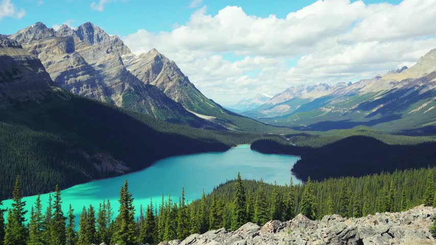 Rocky Mountain peaks rising above a turquoise lake and spruce treetops seen from a rocky viewpoint under puffy clouds, Banff National Park, Alberta, Canada