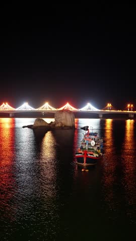 Fishing boat approaching illuminated cable-stayed bridge at night with golden reflections on rippled harbor water and anchored rocky islet nearby, serene maritime scene