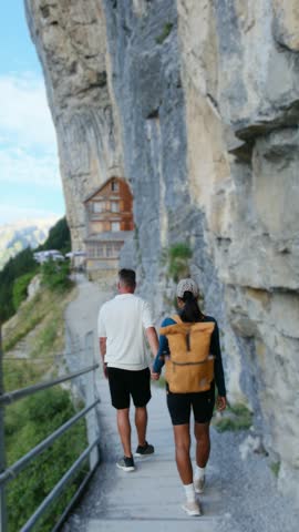 Two hikers follow a narrow cliffside path toward the Wildkirchli mountain inn at Ascher Ebenalp in Switzerland on a bright summer day.