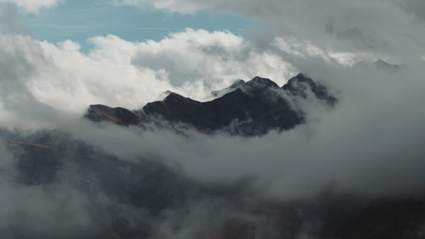 Aerial shot of a rugged alpine mountain massif emerging through thick drifting clouds and mist in Austria, creating a dramatic and atmospheric high mountain landscape.
