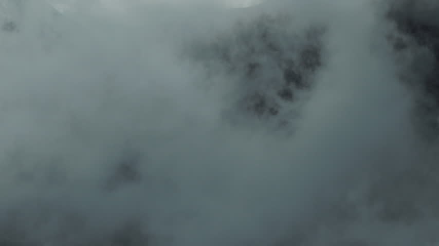 Aerial shot of a rugged alpine mountain massif emerging through thick drifting clouds and mist in Austria, creating a dramatic and atmospheric high mountain landscape.