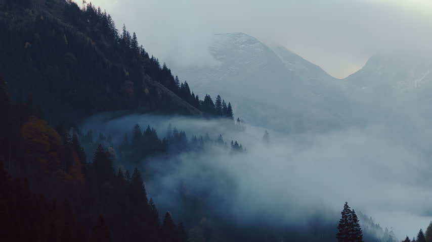 Aerial shot of a rugged alpine mountain massif emerging through thick drifting clouds and mist in Austria, creating a dramatic and atmospheric high mountain landscape.