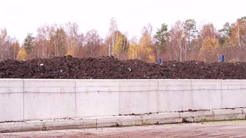 A large mound of dark soil rests behind a concrete retaining wall, with autumn trees and blue posts visible along the roadside