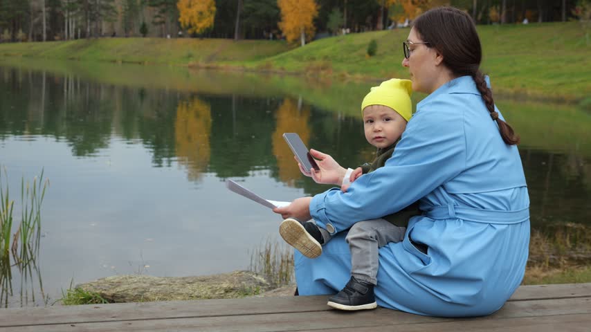 Mother Holds Toddler On Lap While Checking Smartphone And Business Documents Beside Autumn Lake. Busy Female Parent Manages Outdoor Telecommuting Tasks Alongside Curious Child With Calm Focus.