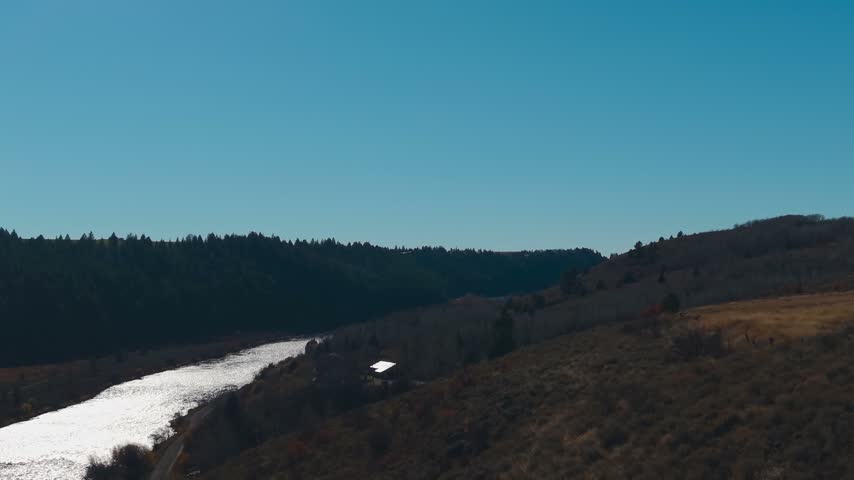 Aerial view following a bird above a sunlit river bordered by wooded banks in Idaho, USA.