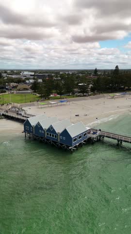 High wide aerial drone shot sweeping past the iconic blue heritage buildings at the start of Busselton Jetty over turquoise water towards the sandy foreshore beach in Australia.