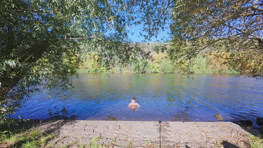 Wide rear view of a shirtless young man swimming away from the shore in a calm lake after cold water immersion in slow motion.