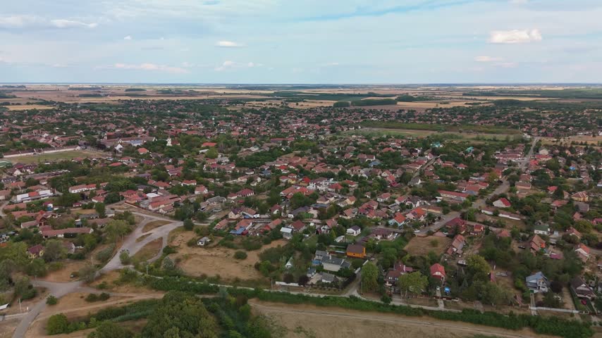 Aerial view of Abádszalók, Hungary, showing a low-rise residential village amid tree-lined streets, open plots and surrounding agricultural fields in summer.