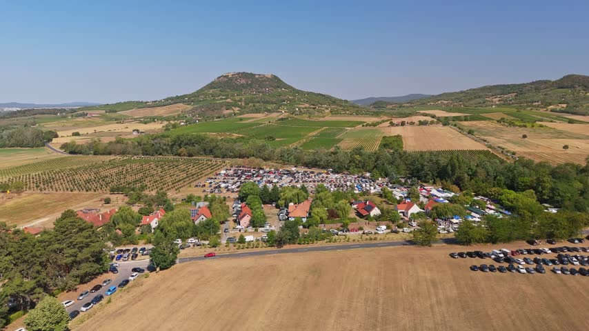 Aerial view of the busy Sunday market and crowded car parks in Káptalantóti, Hungary, surrounded by village homes, orchards, cultivated fields and green hills in summer.