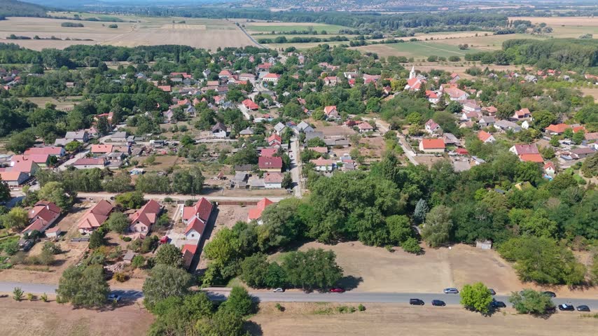 Aerial view of Káptalantóti village in Hungary, showing red-roofed homes, a church, tree-lined streets, cultivated fields and wooded countryside in summer.