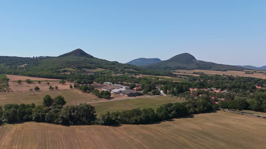 Aerial view of farmland, village homes and agricultural buildings beneath wooded hills in Káptalantóti, Hungary, on a clear summer day.