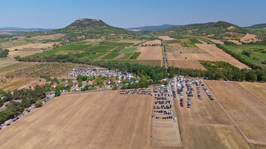 Aerial view of a crowded Sunday market, vendor stalls and large car parks amid farmland and hills in Káptalantóti, Hungary, during summer.
