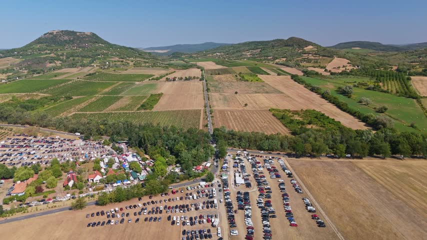 Aerial view of crowded market parking beside farmland and vineyards in Káptalantóti, Hungary, with rolling hills and a busy outdoor market in summer.