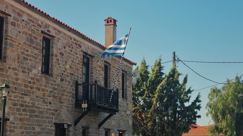 Historic stone building of the Folklore Museum on Ammouliani Island, Halkidiki, Greece, with the Greek flag waving in the breeze under a clear blue sky on a sunny day