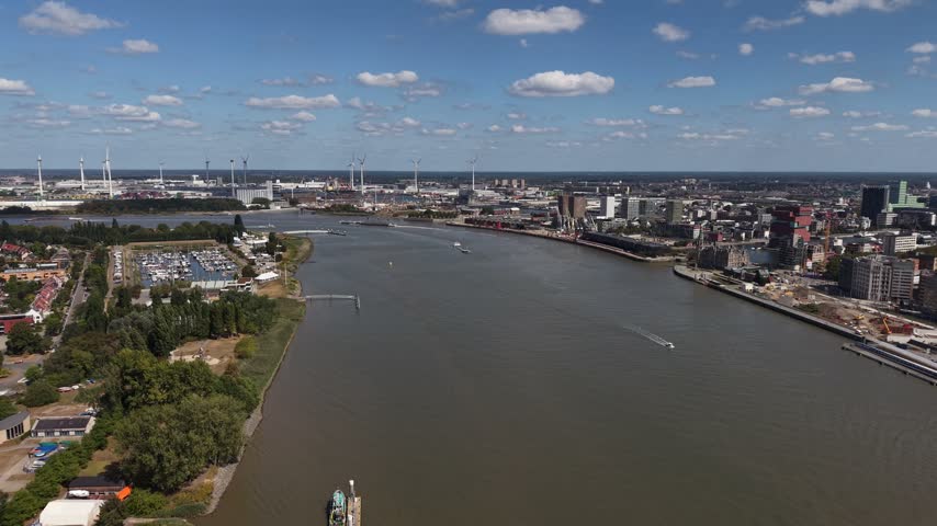 Broad Scheldt River divides Antwerp port skyline from scattered wind turbine fields under blue sky, cargo boats crossing calm water past marina and Belgian riverside homes. Aerial drone wide shot