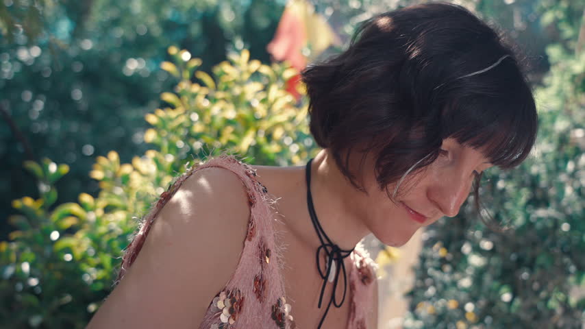 A young woman in a pink sequined dress sits at an outdoor cafe table, leaning her head down as she concentrates on sketching or writing in a notebook.
