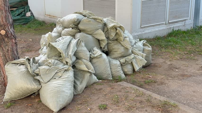 Pile of filled grey polypropylene sandbags stacks on ground near tree trunk and industrial building outdoors. Construction material supply, flood protection barrier or utility engineering debris.