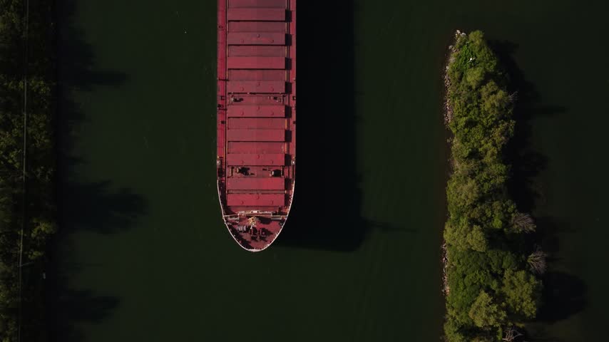 Top-down aerial view of a red cargo ship sailing on dark river water beside a wooded island.