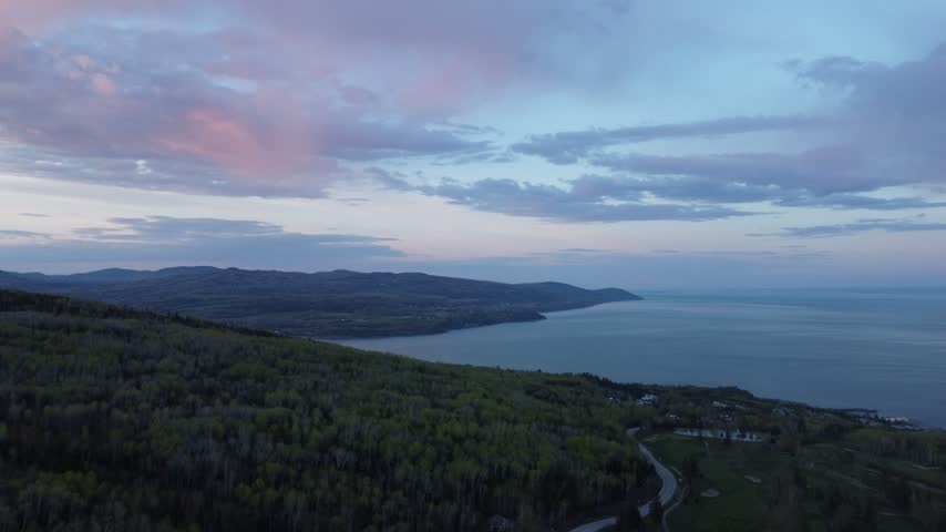 Aerial dusk view over forested hills toward the St. Lawrence River and distant coastline with pink clouds and a winding road below.