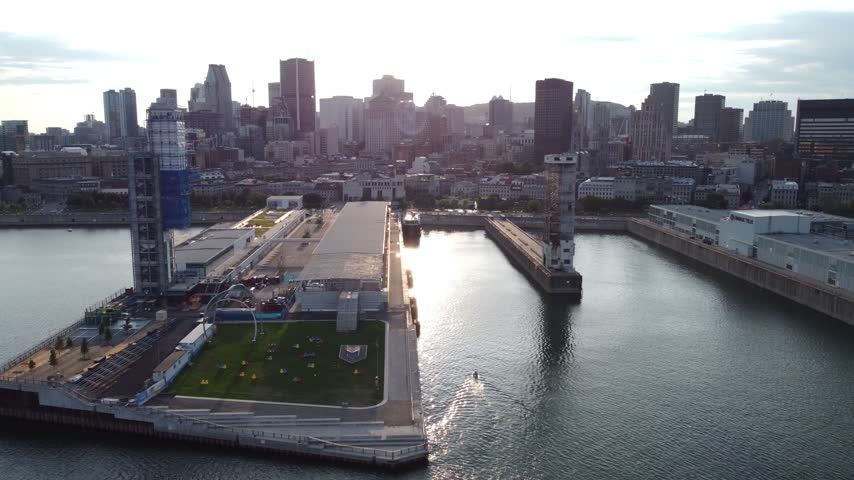 Aerial view of the Old Port of Montreal piers at sunset with downtown skyline silhouettes and sunlight reflecting on the water.