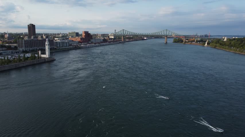 Aerial view above the St. Lawrence River toward the Old Port of Montreal with boats, marina, clock tower and bridge in the distance at dusk.