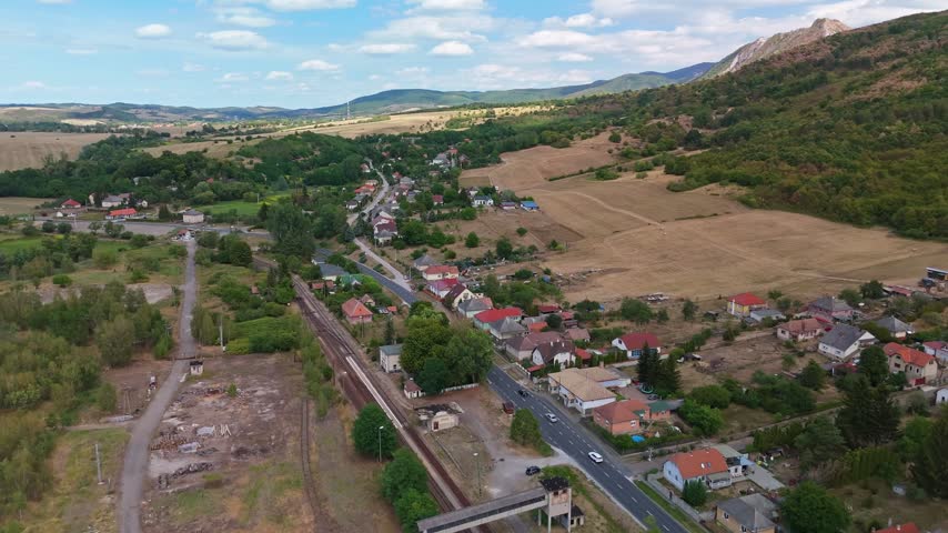 Aerial view of railway tracks and homes in Mónosbél, Hungary, surrounded by harvested fields, forested slopes and the Bükk Mountains on a summer day.