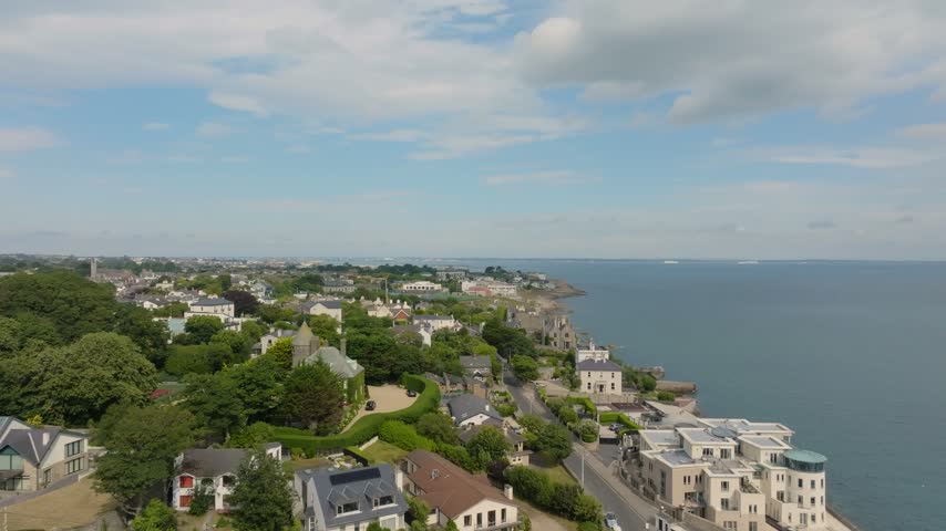 An aerial view follows a rocky coastline past waterfront houses, a turreted stone building and sports courts beside the sea. Dalkey Co.Dublin