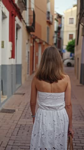 A woman walks down a narrow street in a small town. She wears a white dress and carries a bag. The sun shines on the buildings and the pavement.