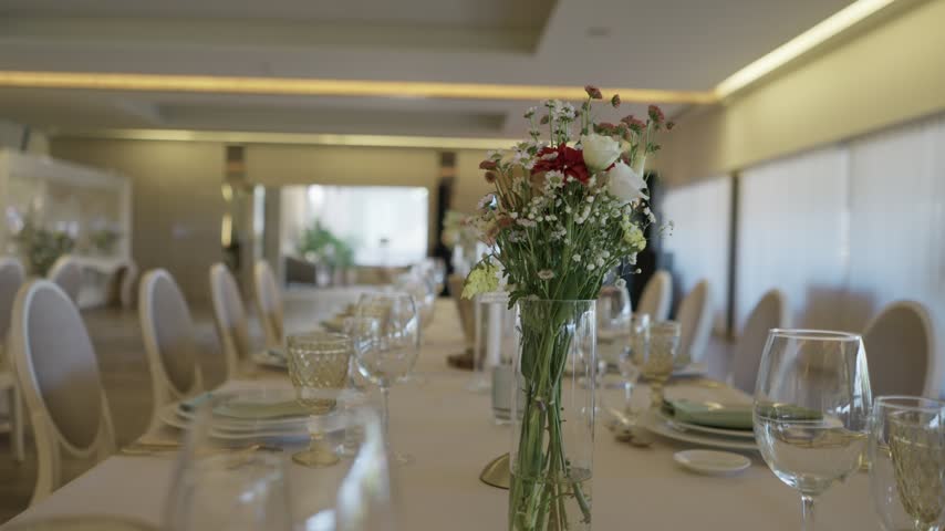 Long banquet table set for a wedding with a glass vase of wildflowers, wine glasses, plates and white chairs in a large reception hall.
