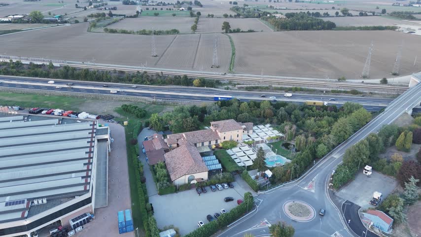 Villa Costanza with terracotta roof, courtyard, glass conservatory and blue pool beside warehouse and A1 motorway, Pontenure, province of Piacenza, Emilia Romagna, Italy, drone forward flyover descent