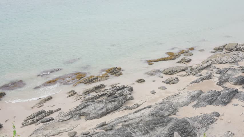 Slow motion layered rock strata on sandy beach with clear shallow sea from above