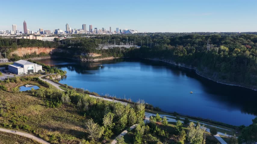 Bellwood Quarry reservoir at Westside Park in Atlanta, Georgia reflects blue sky below rock cliffs, with a white water works building, trail and the downtown skyline beyond. Aerial drone wide shot