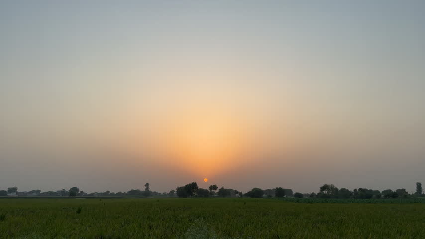 The sun sets over a green agricultural field with trees along the horizon.