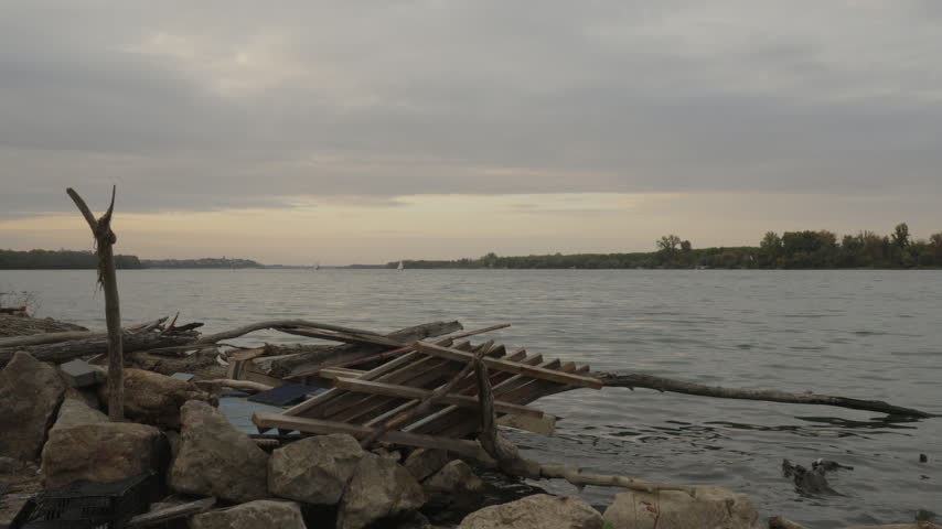 Wide river landscape at dusk with distant birds flying over calm water beneath a cloudy evening sky. Driftwood, fallen tree trunks, rocks and discarded wooden structures lie along the rocky shoreline. Muted colors create a quiet, atmospheric natural scene.