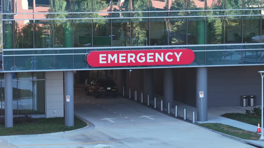 Closer view of the red EMERGENCY sign over the covered drive-in entrance of a hospital emergency department with bollards, cones and a parked SUV in Atlanta, Georgia. Aerial view