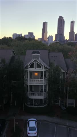 Single lit window glows in a tall apartment building with stacked balconies below the downtown Atlanta skyline at twilight, as trees ring the parking lot in Georgia. Aerial view