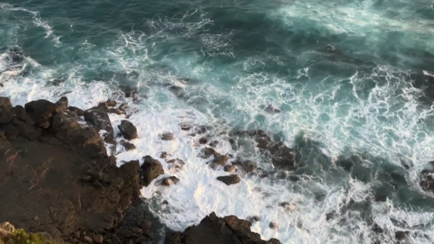 High angle view of turquoise ocean waves crashing with white foam onto a rocky coast at golden hour